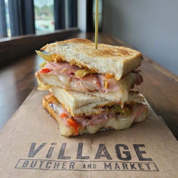 A grilled sandwich with melted cheese, ham, and peppers on a branded Village Butcher and Market paper.