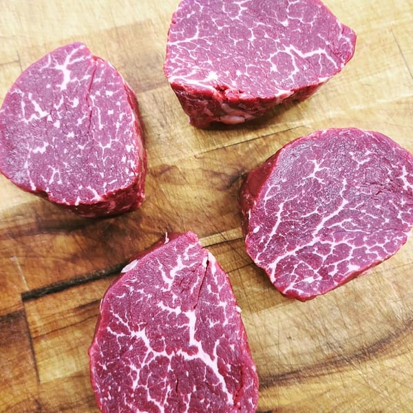 Four raw, marbled cuts of beef, likely filet mignon, on a wooden cutting board.