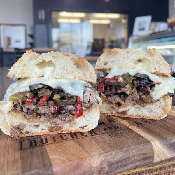 Two halves of a Village Butcher and Market cheesesteak sandwich with melted cheese, peppers, and mushrooms.